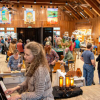 A bustling crafts fair with visitors browsing tables, colorful banners, and a person playing the piano.