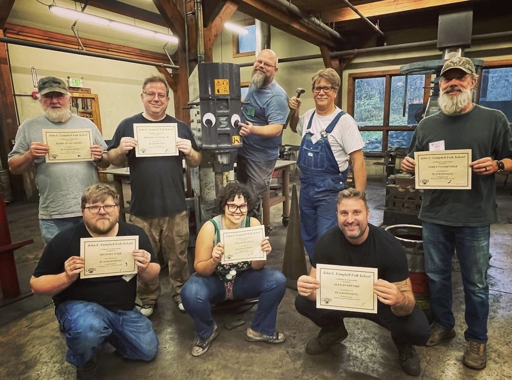 Eight students posing and smiling in the Blacksmithing shop, holding up their certificates of completion