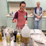 The instructor, a young woman, points towards a table of ingredients and is explaining the process to students.