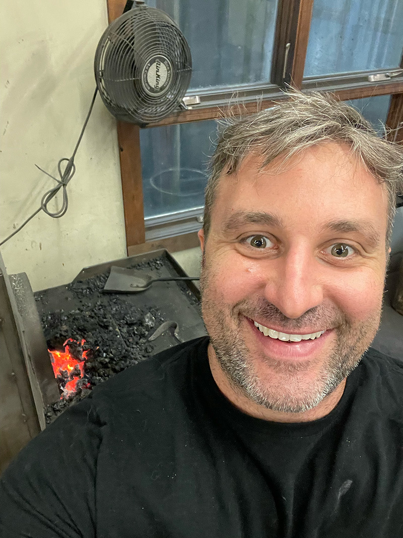 Alex smiles with his blacksmithing forge in the background.