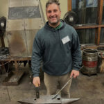 A young man smiling at the camera, standing at his forge with hammer and steel in hand