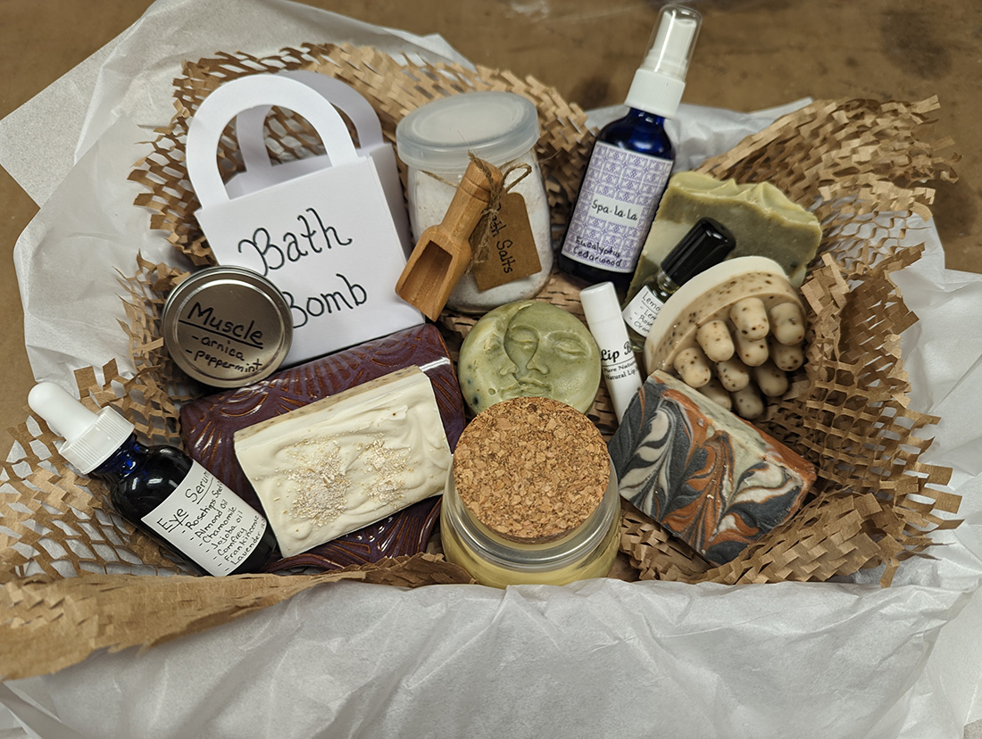A basket full of handmade soap and other handmade creations, with handwritten labels. Objects include a bar of soap, a jar of eye serum, lip balm, "Spa-la-la" Eucalyptus spray, bath bombs, and more.
