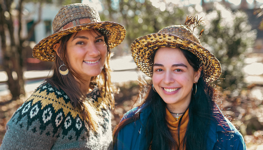 Two students stand in a woodsy area wearing their handmade willow basket hats
