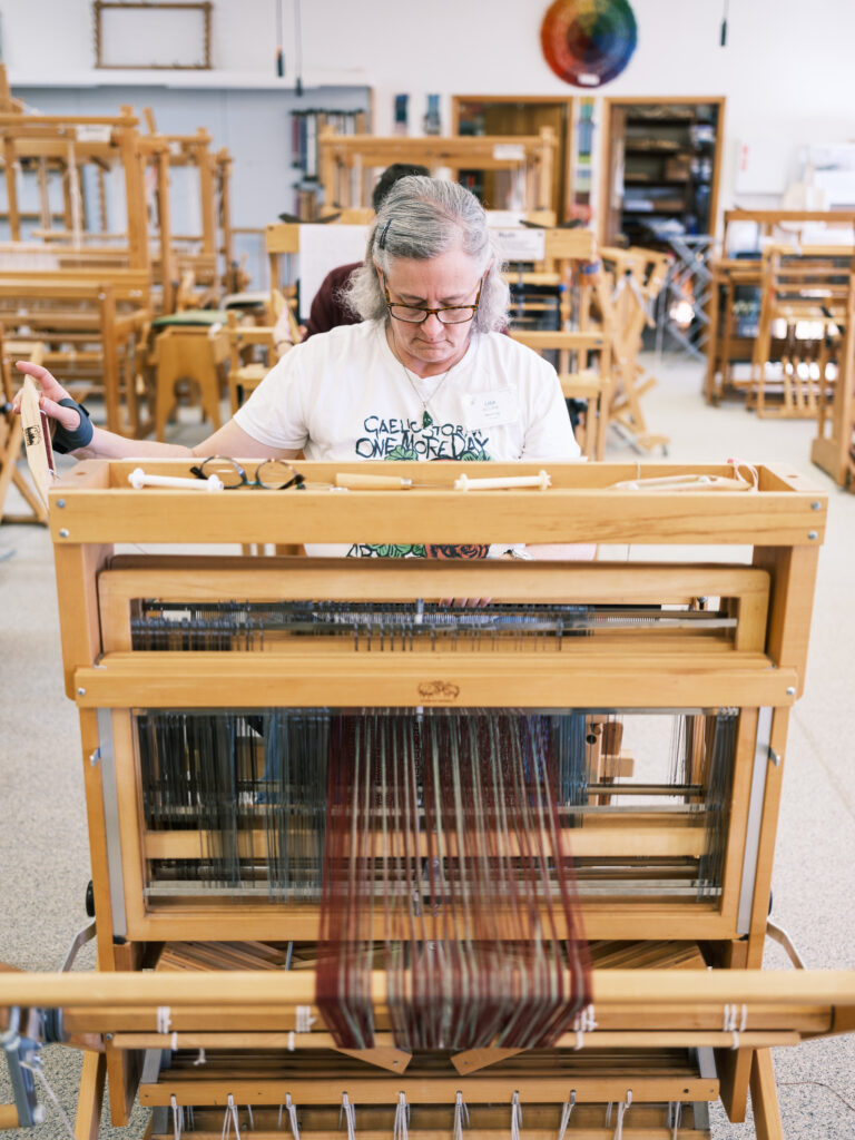 2024-02-04-Weaving-Sashiko-Ori-Beth-Ross-Johnson--and-Phyllis-Miller-3 A person is focusing on weaving at a loom in what appears to be a workshop or class setting.