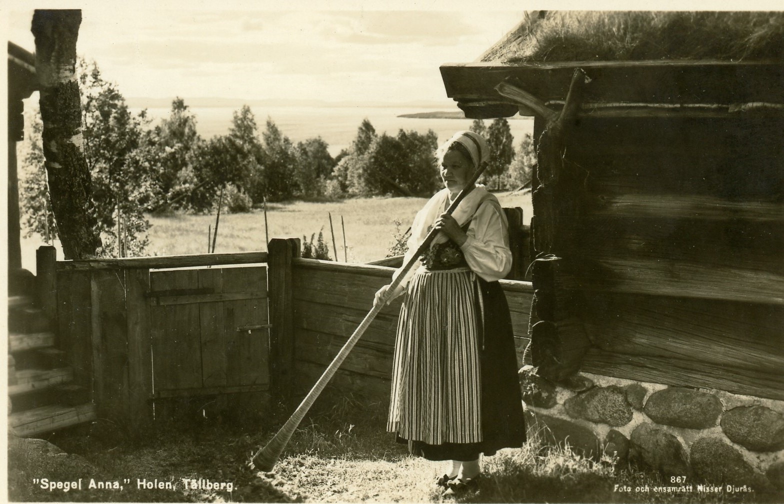 black-and-white postcard photograph of a woman wearing traditional Scandinavian dress working in a yard next to a log and stone building