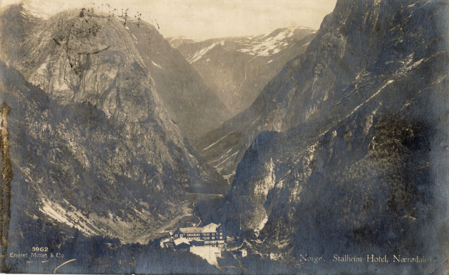 black-and-white postcard photograph of enormous mountains dwarfing a multi-story inn building in the center of a small valley