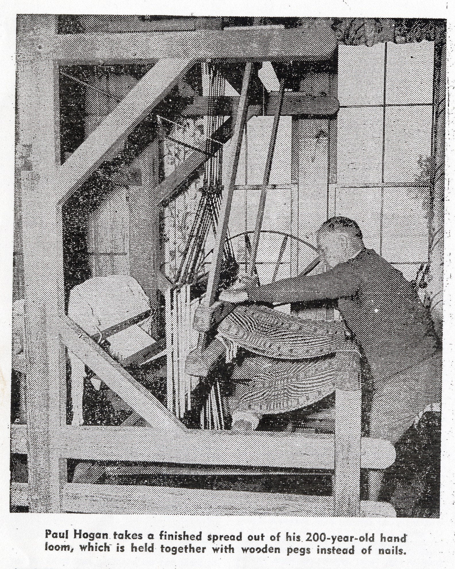 Hogan01 newspaper photo of a man working on a coverlet on a loom, captioned "Paul Hogan takes a finished spread out of his 200-year-old hand loom, which is held together with wooden pegs instead of nails."