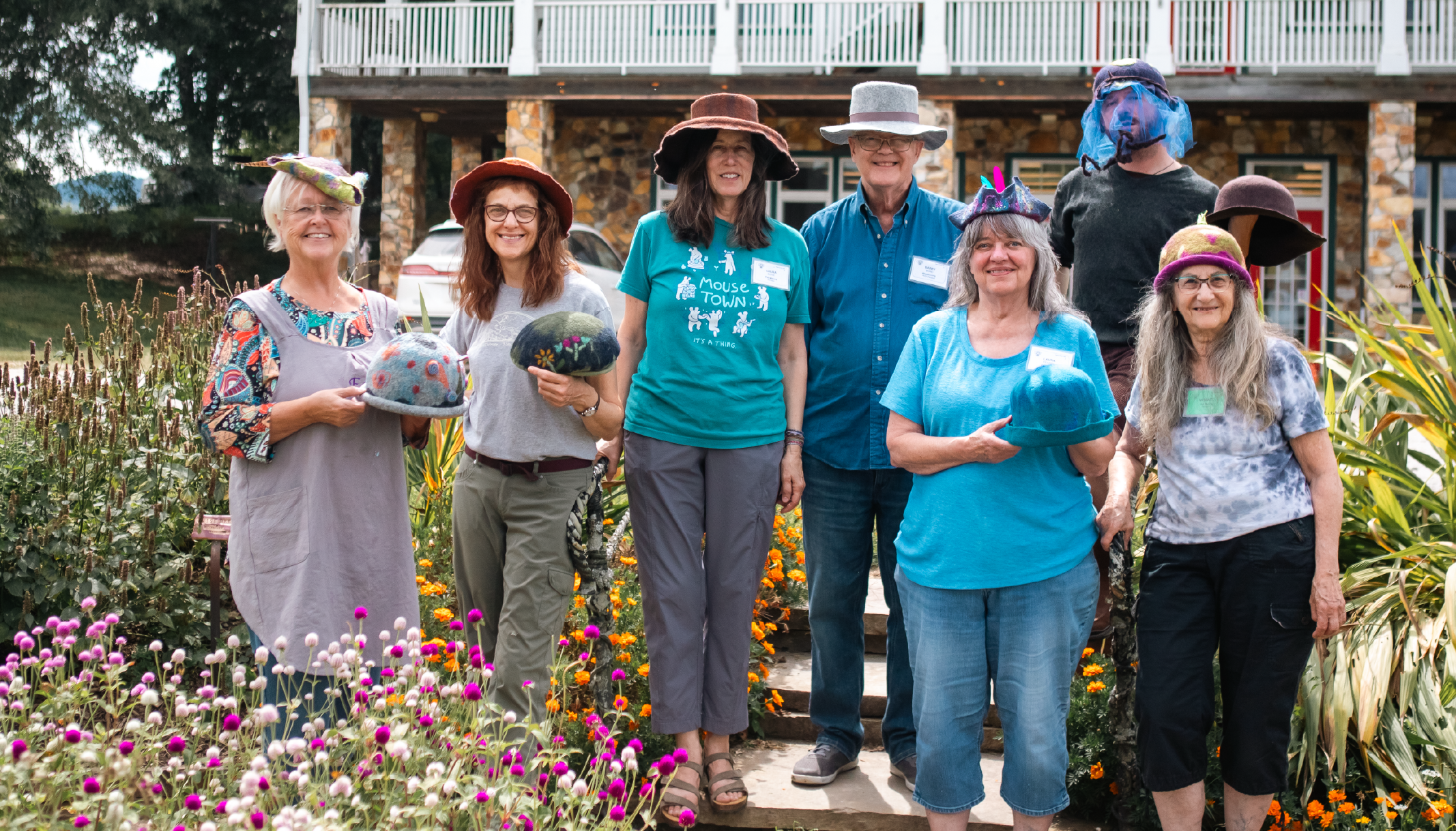 A group of people stand outside, wearing colorful hats, smiling, with flowers in the foreground.