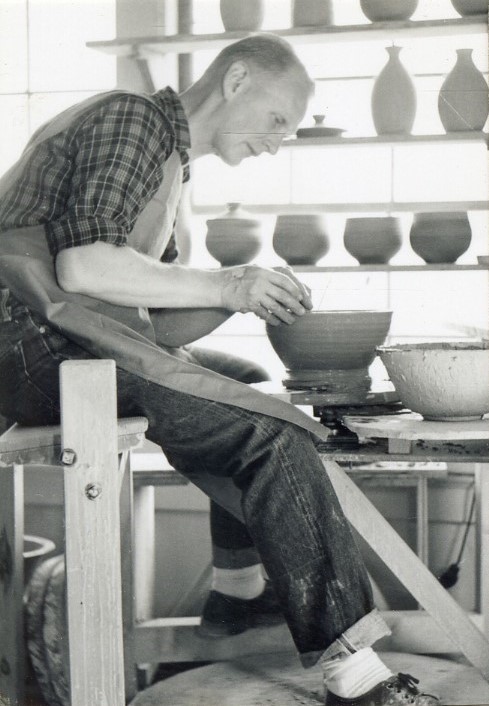 black-and-white photograph of a blonde man wearing jeans and a plaid shirt sitting at a potter's wheel and making a clay bowl