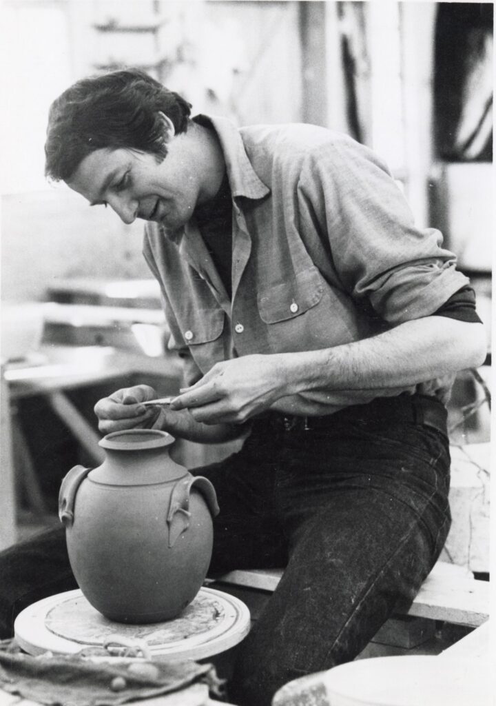 black-and-white photograph of a dark-haired man wearing dark jeans and a light-colored button-down shirt making a two-handled ceramic vase on a potter's wheel