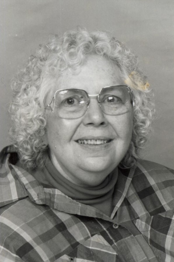 black-and-white photograph of a woman with shoulder-length curly white hair and glasses, wearing a plaid shirt over a turtleneck