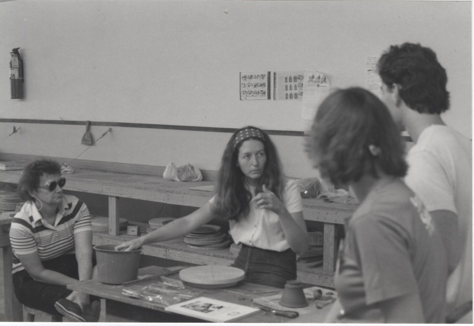 black-and-white photograph of a woman with long hair and a headband teaching three other people in a pottery studio