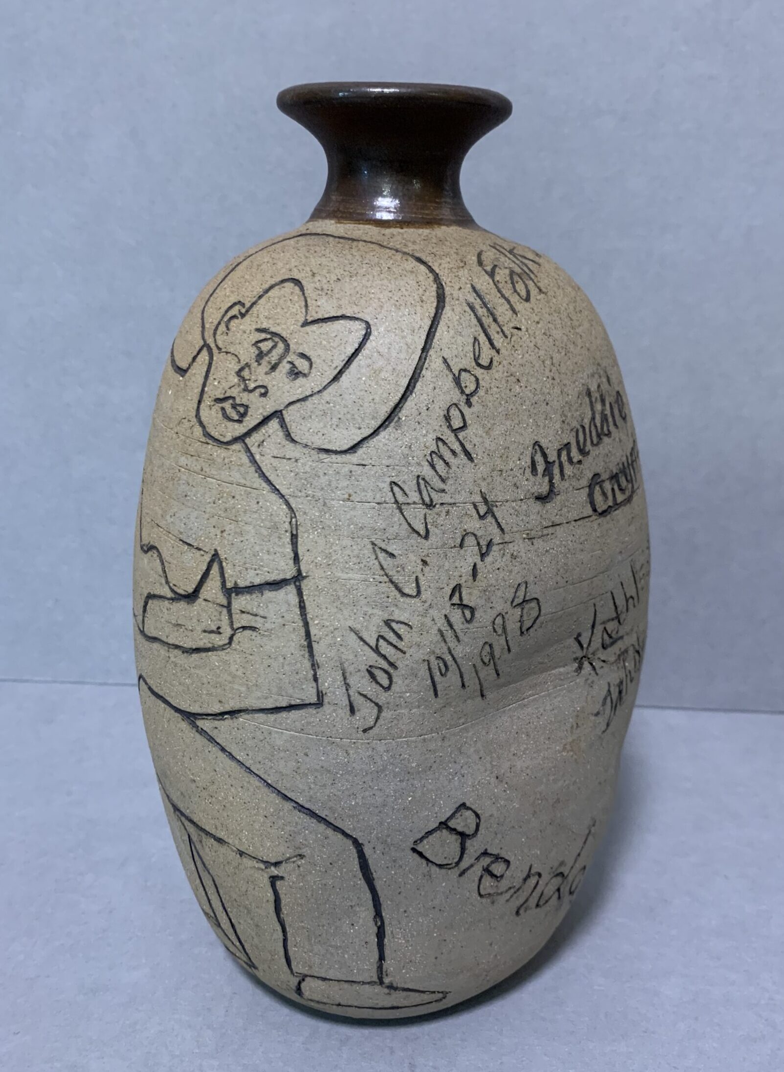 tall, round, unglazed ceramic vase with brown glazed spout on top; sides etched with image of a female potter at a wheel and text "John C. Campbell Folk School 10/18-24 1998" and several names