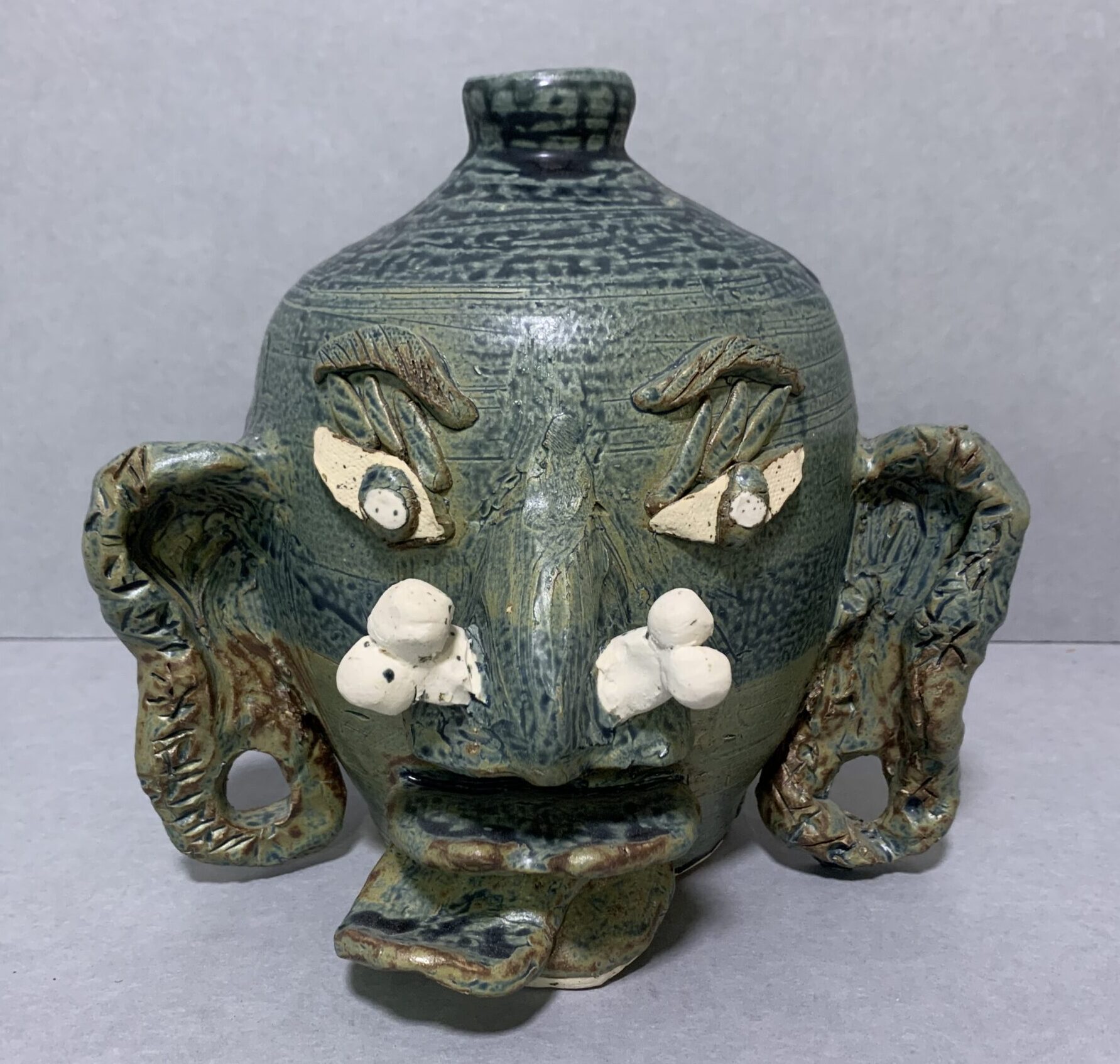 blue-gray glazed ceramic face jug with oversized ears with gauge holes, an unglazed ceramic bone sticking through the nose, and protruding tongue