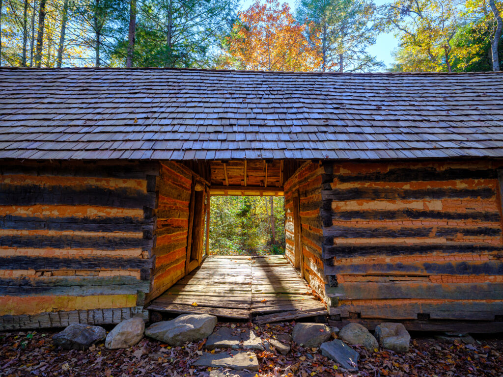 A rustic wooden cabin with an open passage, surrounded by trees with autumn foliage.