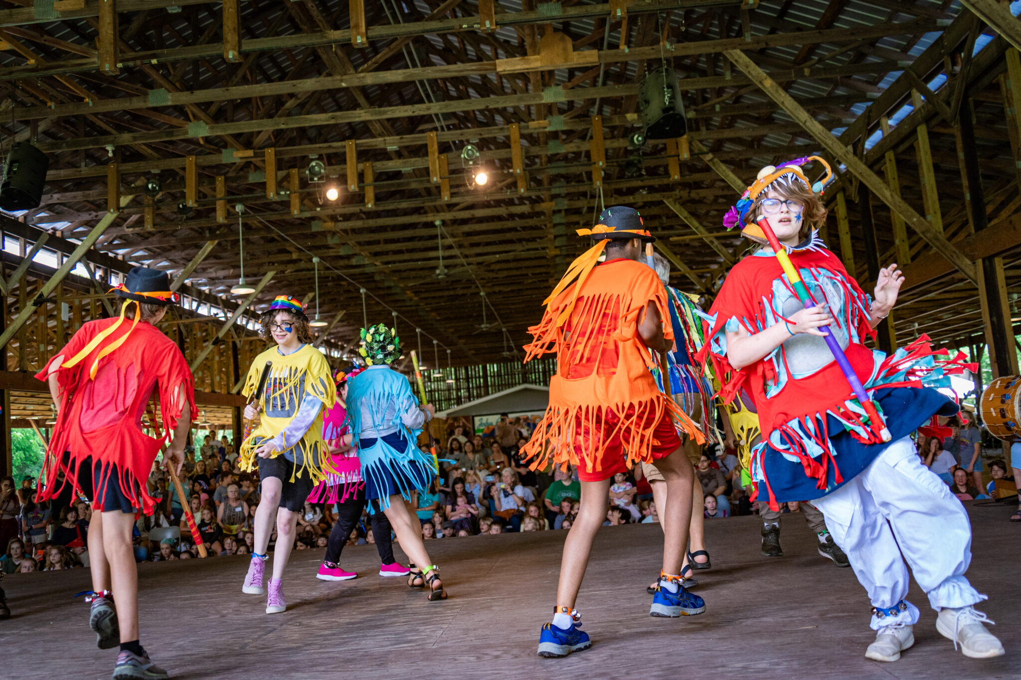 People in colorful costumes are performing a lively folk dance on a wooden stage, watched by a seated audience.
