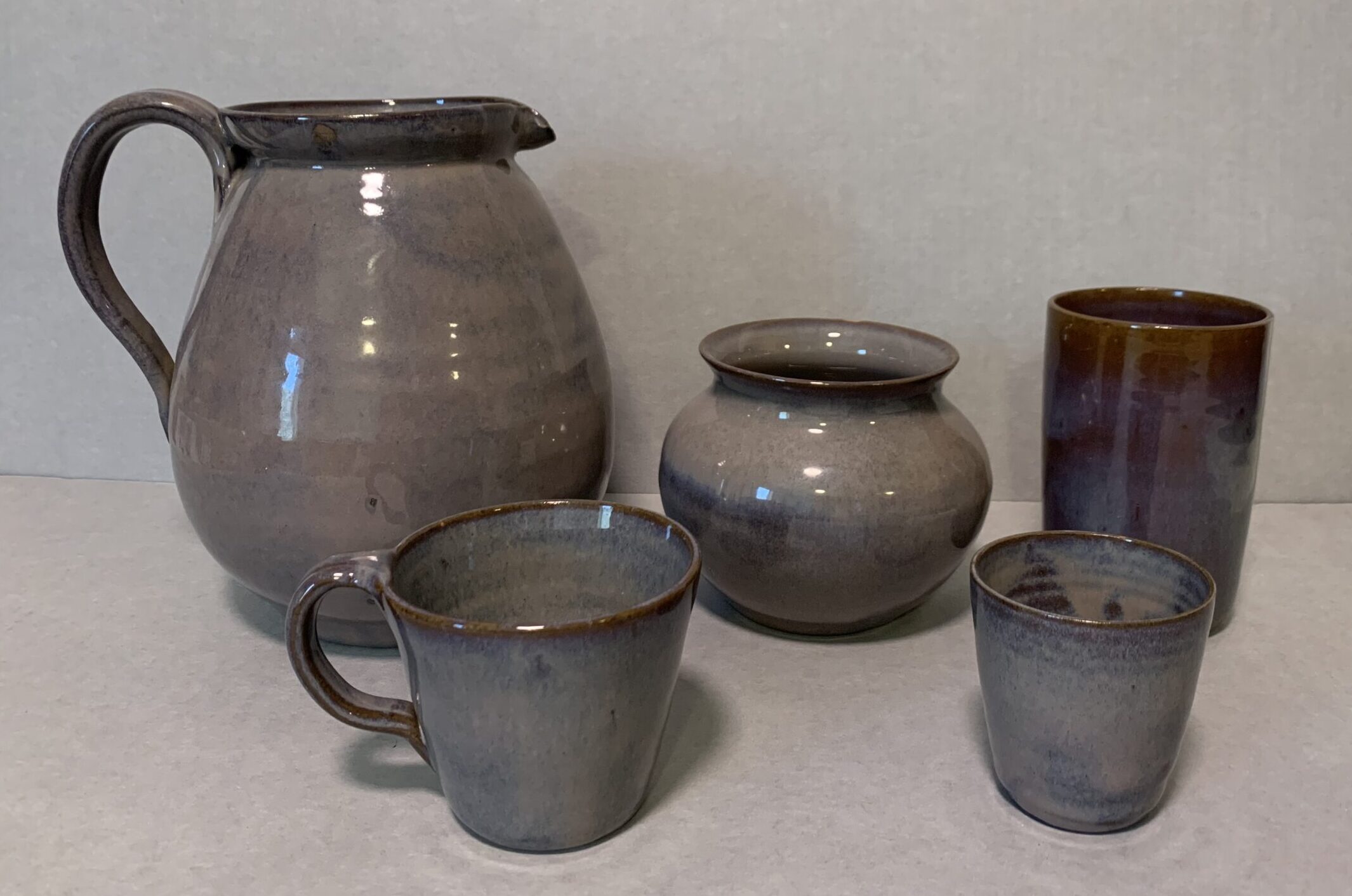 set of purple-ish and brown glazed ceramic dishes including a pitcher, wide-mouthed vase or jar, tumbler, short drinking cup, and mug