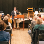 An auction event is taking place with attendees seated and a person speaking at a podium labeled "Folk School."