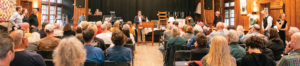 An auction event is taking place with attendees seated and a person speaking at a podium labeled "Folk School."