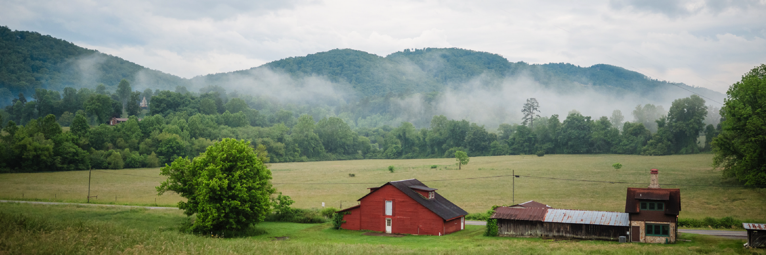 A rural landscape features red barn-like structures, open fields, lush greenery, and misty hills.