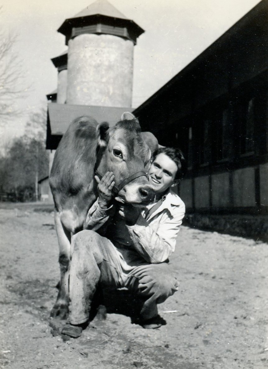 2024-14-313_cropped black-and-white photo of a smiling man crouched next to a cow and gently holding her face, with barn and silos in background